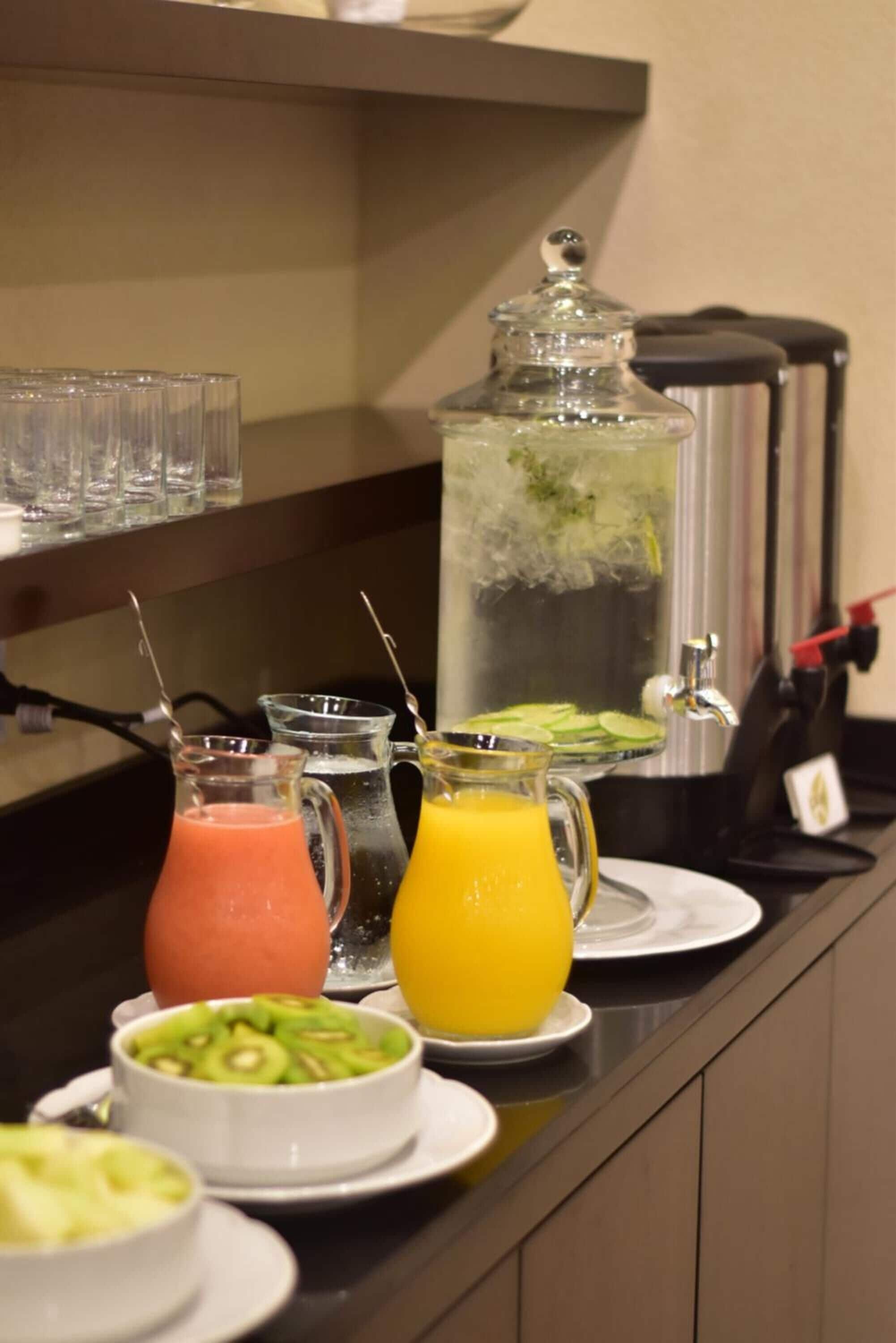 A breakfast buffet with various drinks, a large pitcher, and sliced fruits on a counter.