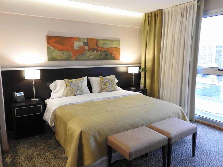 A hotel room with a large bed, two bedside tables with lamps, a bench, a painting on the wall, and a window with curtains.