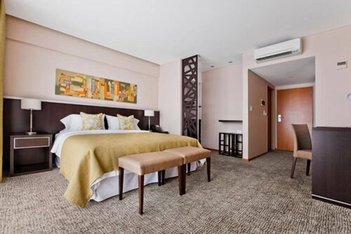A hotel room with a bed, a bench, a desk, and an air conditioning unit.