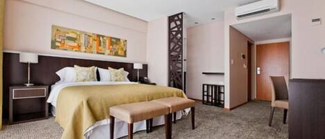 A hotel room with a bed, a bench, a desk, and an air conditioning unit.