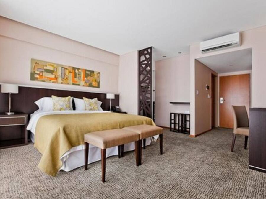A hotel room with a bed, a bench, a desk, and an air conditioning unit.