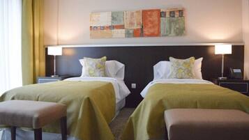 A hotel room with two beds, each with a green bedspread and matching pillows. There is a nightstand with a lamp on each side of the beds. A large abstract painting is on the wall above the beds.
