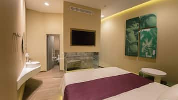 A modern hotel room with a large bed, a built-in TV, a small round table, and a bathroom with a glass shower enclosure.