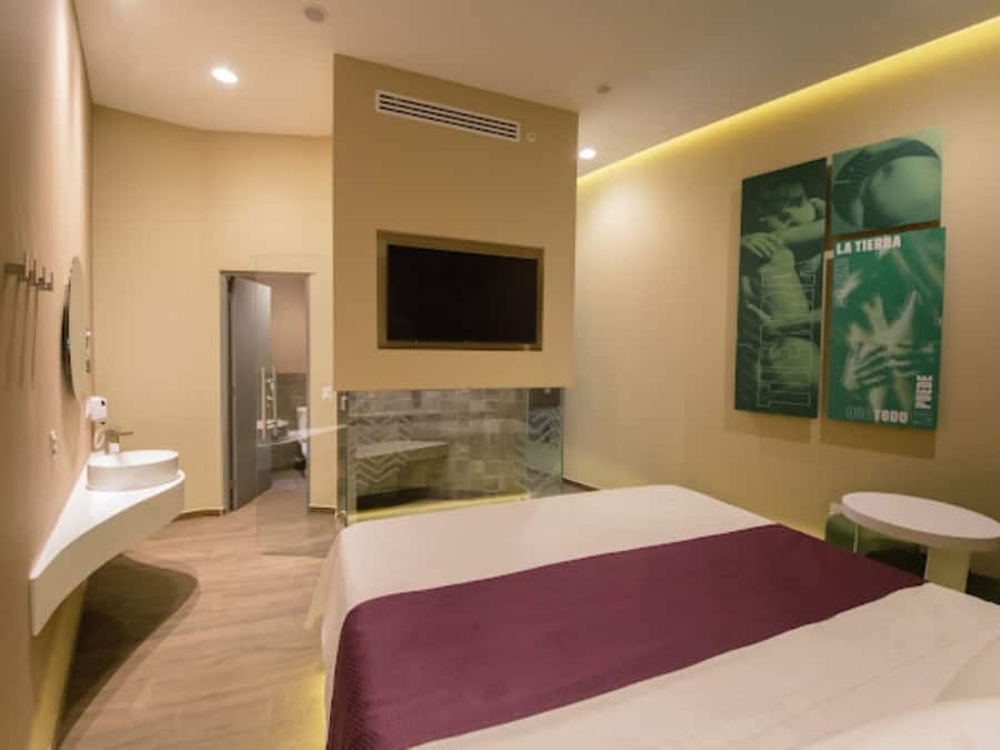 Una habitación de hotel moderna con una cama grande, un televisor integrado, una mesita redonda pequeña y un baño con cabina de ducha de vidrio.