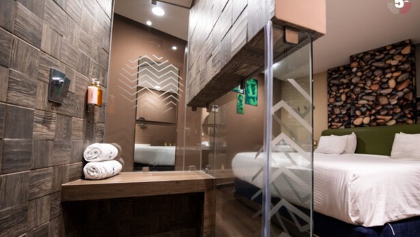 Une chambre d’hôtel moderne avec un mur en bois, une douche en verre, un lit avec du linge de maison blanc et un banc en bois.