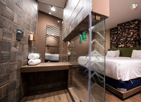 Una habitación de hotel moderna con una pared de madera, un box de ducha de vidrio, una cama con ropa blanca y un banco de madera.