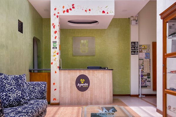 A reception area with a wooden desk, a couch, and a wall-mounted sign for Ayenda Hoteles. - Ayenda 1140 Roma Plaza (Manizales)