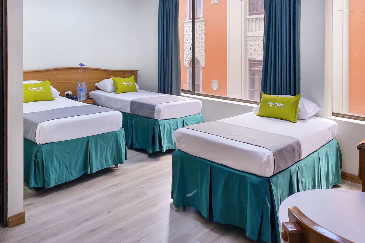 A hotel room with two beds, each with a green bed skirt and a pillow labeled "Ayenda Hotels".