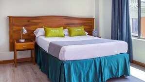 Un lit bien fait, avec une tête de lit en bois, deux oreillers verts, un oreiller blanc et un couvre-lit turquoise. Une table de chevet en bois, sur laquelle se trouvent une lampe et une bouteille d’eau, est placée à côté du lit.