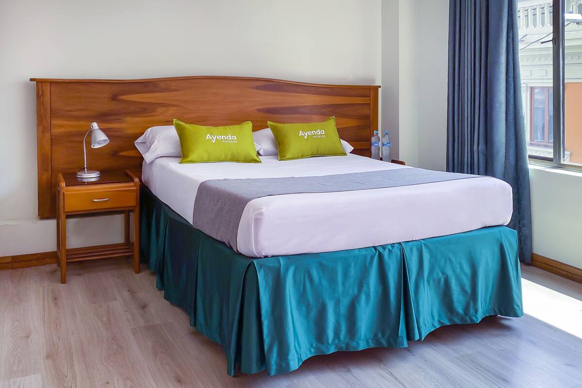 A neatly made bed with a wooden headboard, two green pillows, a white pillow, and a teal bed skirt. There is a wooden nightstand with a lamp and a bottle of water beside the bed.