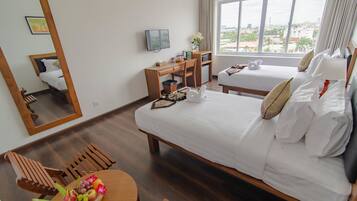 Una habitación de hotel con una cama grande, un escritorio con computadora, una mesita con fruta y flores, y un ventanal con vista a la ciudad.