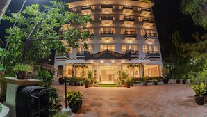 Un hotel de varios pisos con una entrada central, rodeado de plantas en macetas y un patio empedrado.