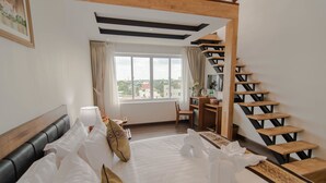 Habitación de hotel con cama, escritorio, silla, escalera y ventana con vista a edificios.