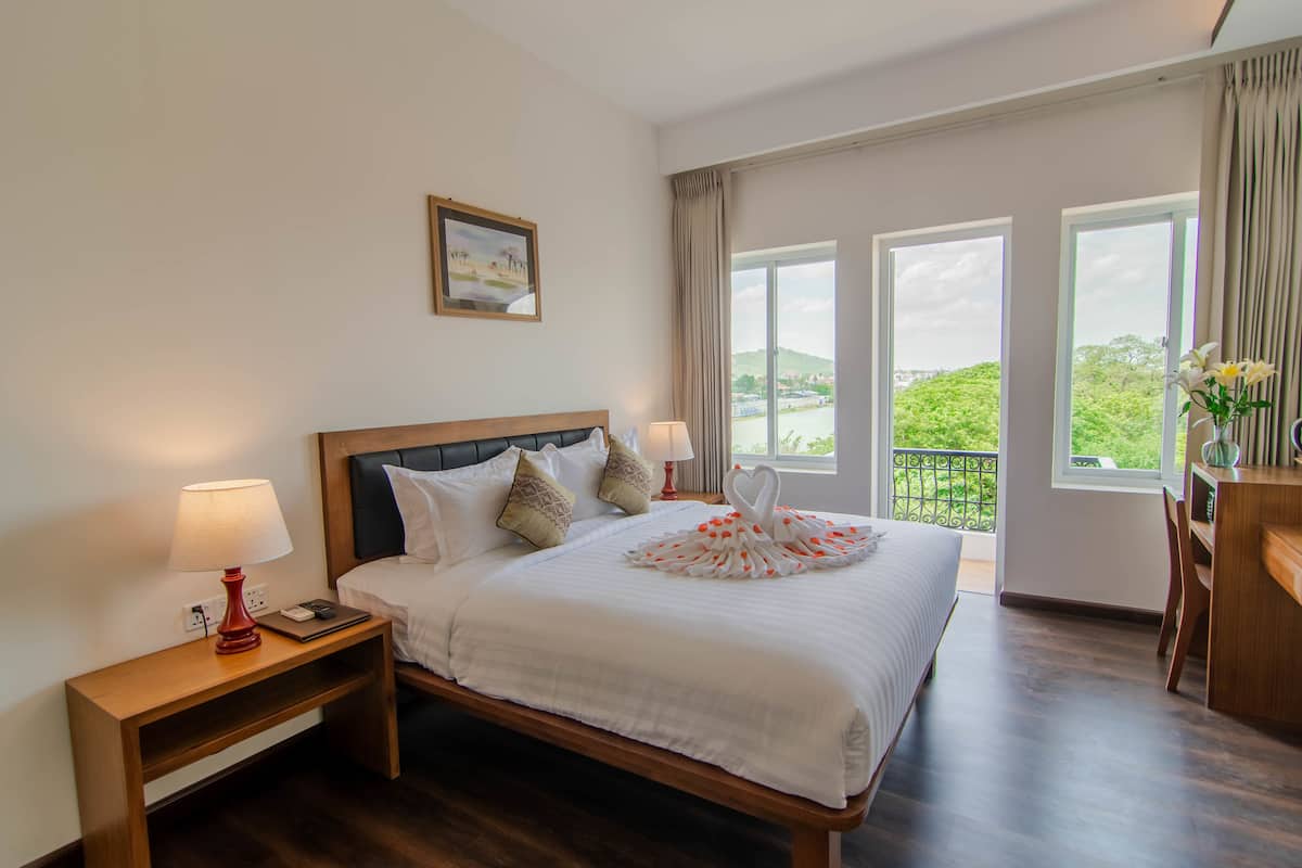 A hotel room with a large bed, two bedside tables, a desk, and a view of greenery through the windows.