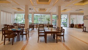 Un comedor amplio con varias mesas y sillas, ventanas grandes con persianas y plantas de interior.