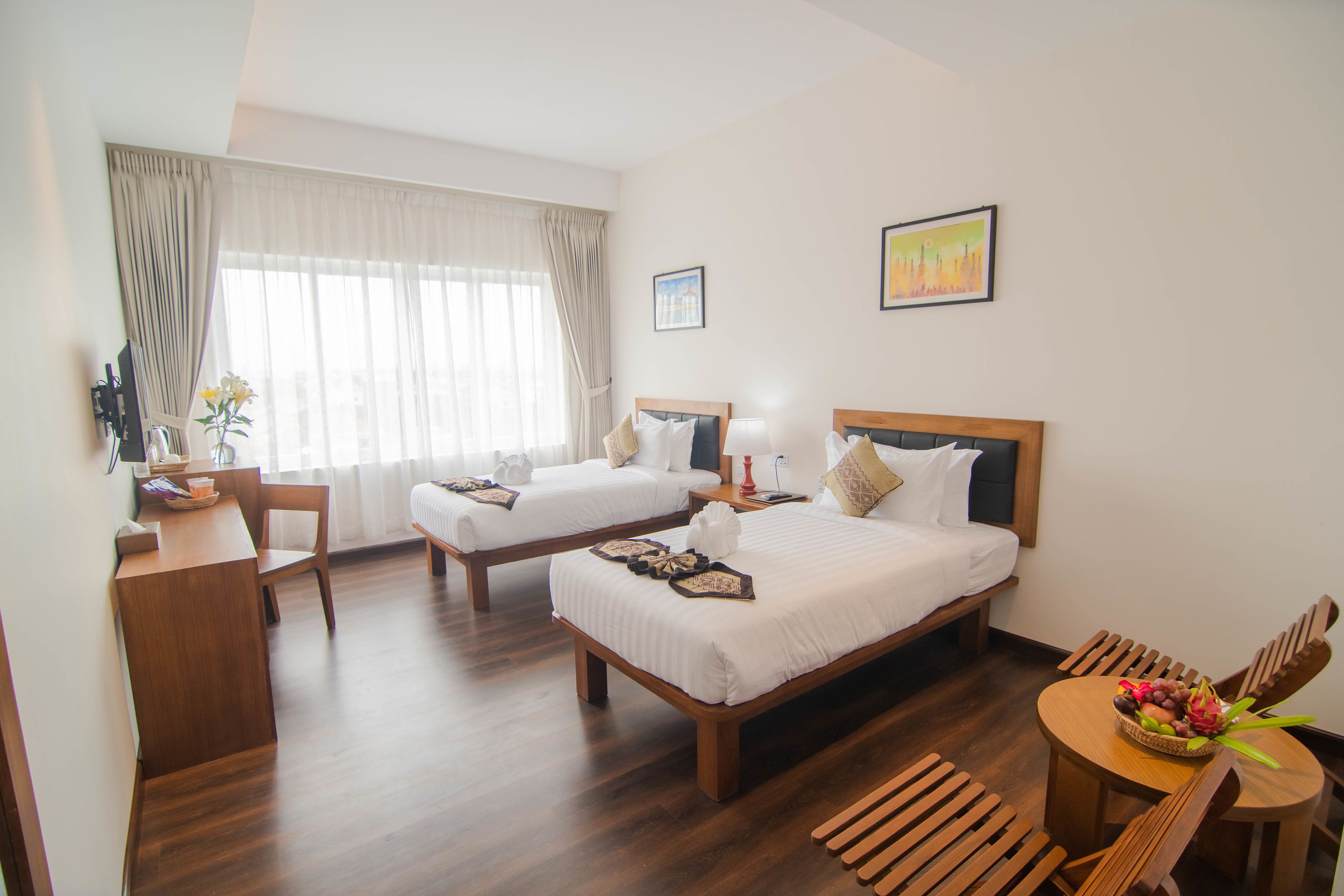 A hotel room with two beds, a desk, a chair, a small table with a fruit bowl, and a window with curtains.