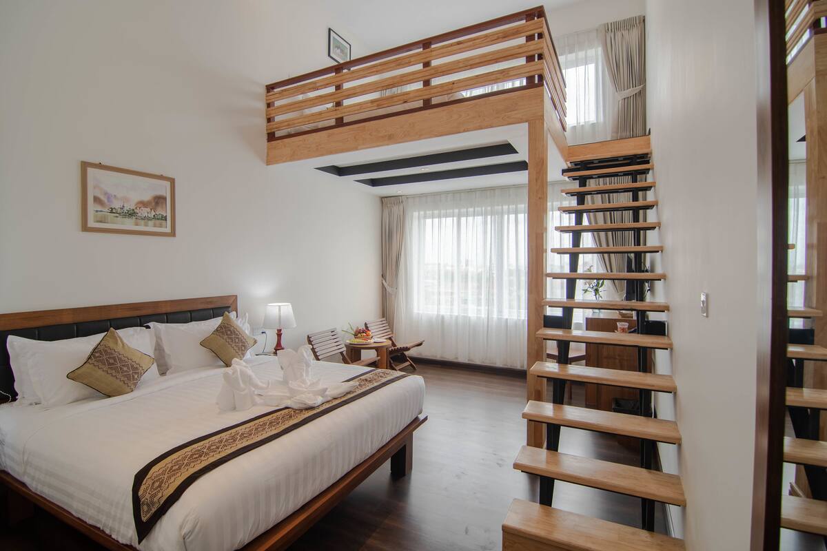 A bedroom with a wooden bunk bed, a large bed with white linens, a bedside table with a lamp, and a window with curtains.
