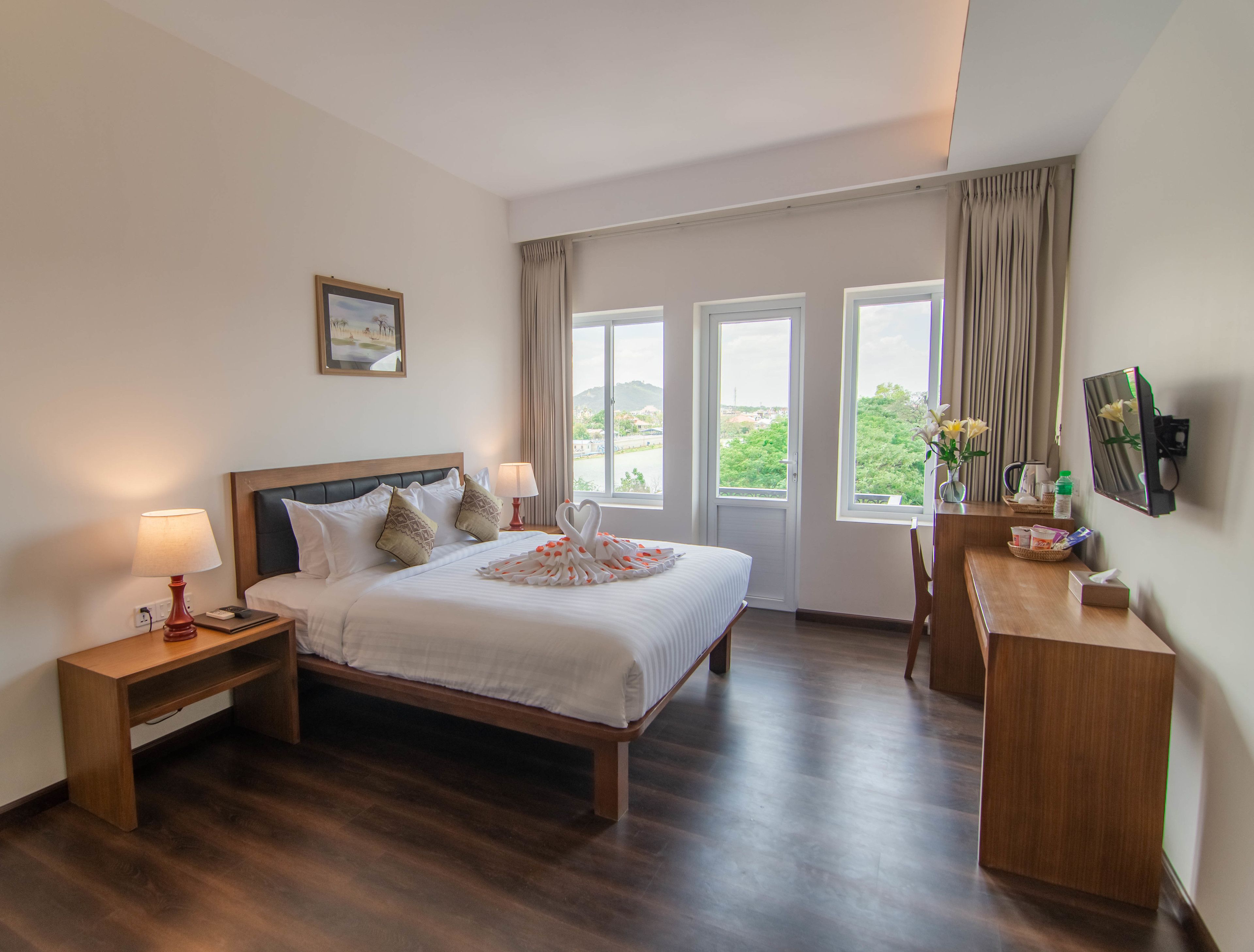 A hotel room with a large bed, two bedside lamps, a wooden desk, and a view of greenery through the windows.