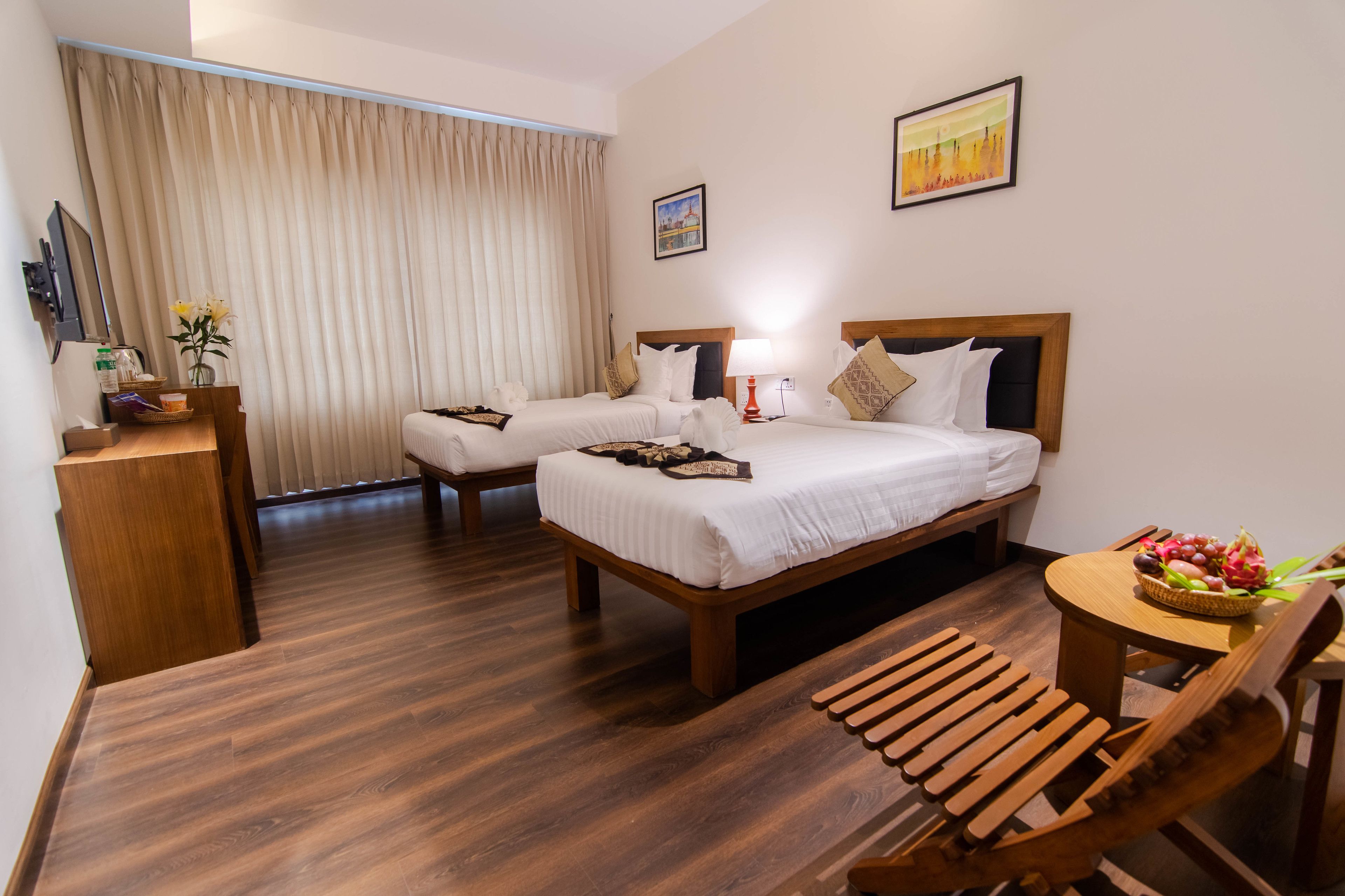 Una habitación de hotel con dos camas, un escritorio de madera, una silla, una mesita con una cesta de fruta y un cuadro en la pared.