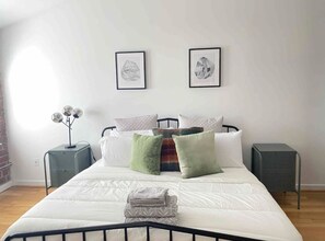 Een slaapkamer met een bed, twee nachtkastjes, een lamp en twee ingelijste foto’s aan de muur.