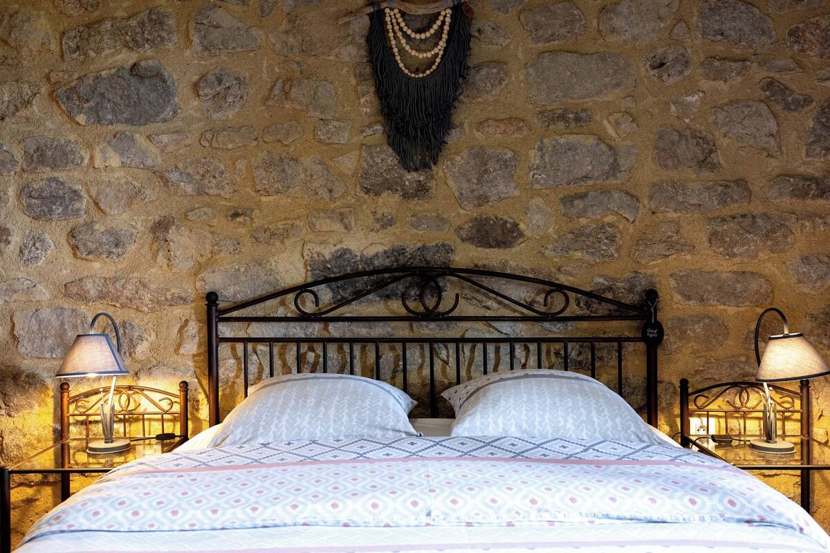 A bed with a patterned bedspread, two bedside lamps, and a stone wall with a hanging decoration.
