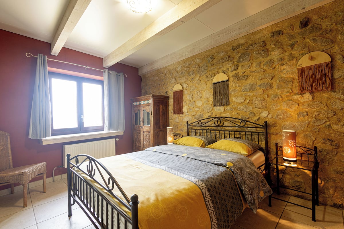 A bedroom with a stone wall, a bed with patterned bedding, a chair, and a bedside lamp.