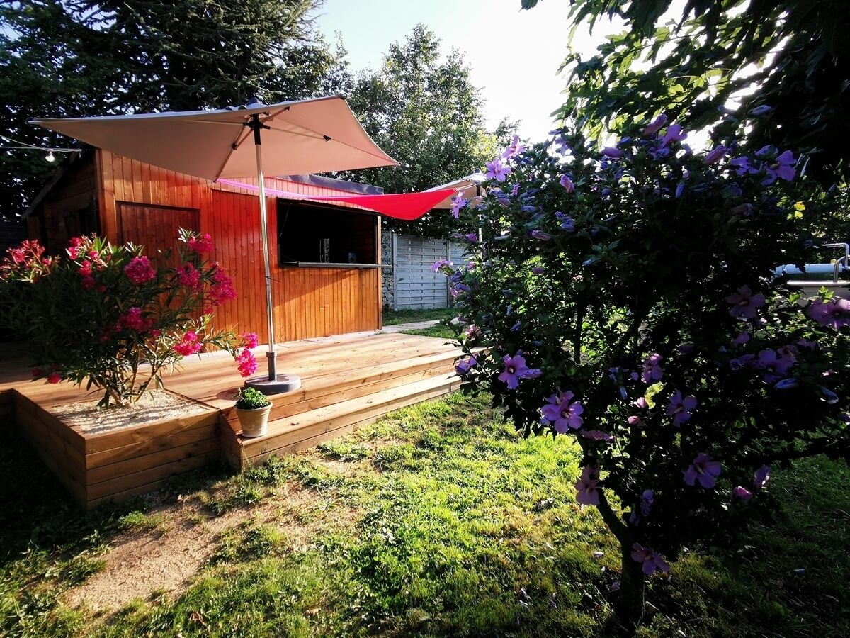 Una terraza de madera con un toldo rojo, rodeada de plantas y árboles en flor.