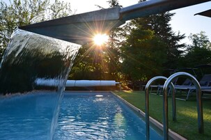 Una piscina con una cascada, rodeada de césped y sillas de descanso.