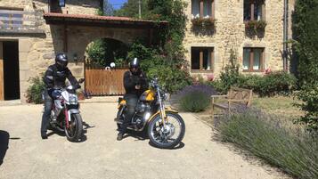 Two motorcycles parked in front of a stone building with a wooden gate and a bench in the foreground.