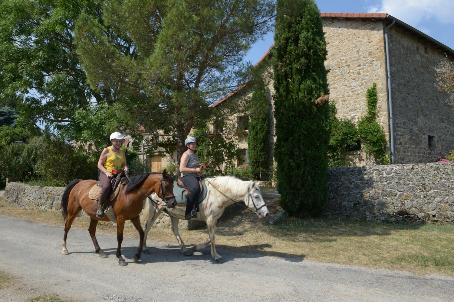 Dos personas montando a caballo por un camino al lado de un edificio de piedra con techo rojo.