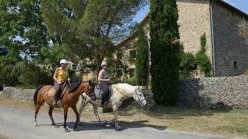 Dos personas montando a caballo por un camino al lado de un edificio de piedra con techo rojo.
