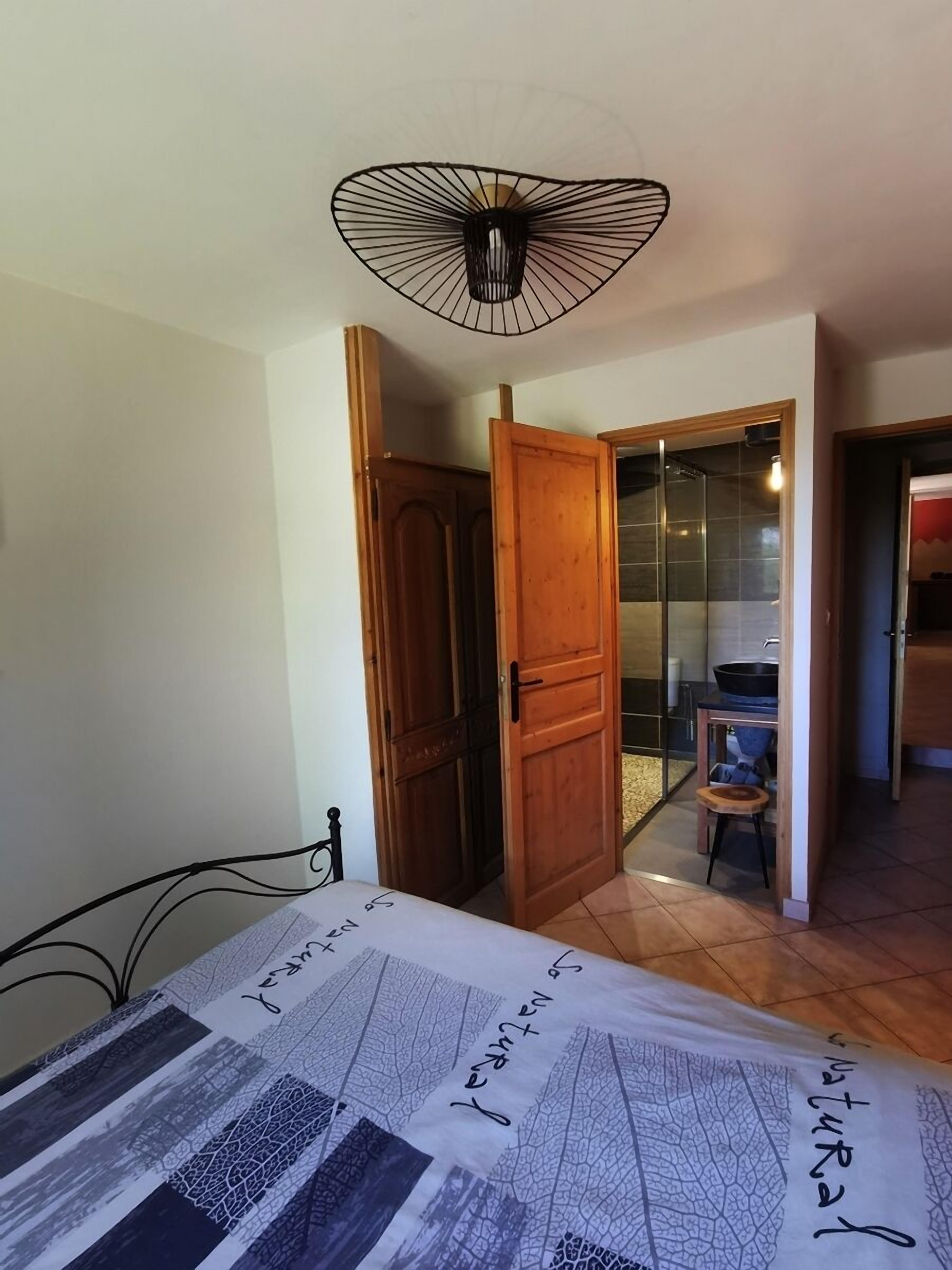 A room with a bed, a wooden door, a small stool, and a kitchen area visible through an open door.