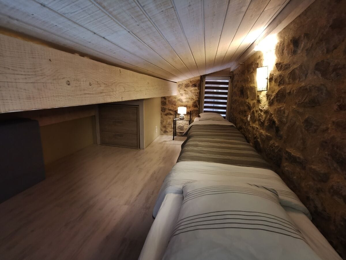 Una habitación estrecha y tenuemente iluminada, con techo inclinado y paredes de piedra. Una cama con colcha a rayas está ubicada contra la pared derecha, y una mesita de noche con lámpara cerca del centro.