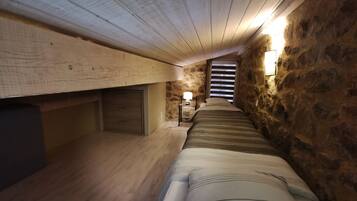 A narrow, dimly lit room with a sloped ceiling and stone walls. A bed with a striped blanket is positioned against the right wall, and a small table with a lamp is near the center.