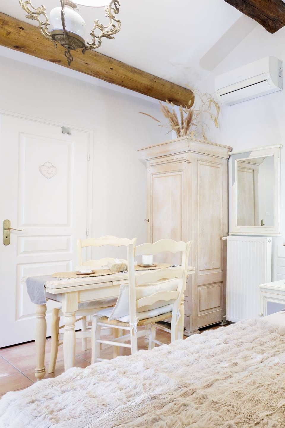 A room with a wooden ceiling, a white wardrobe, a white chair, a small table, and a fluffy rug.