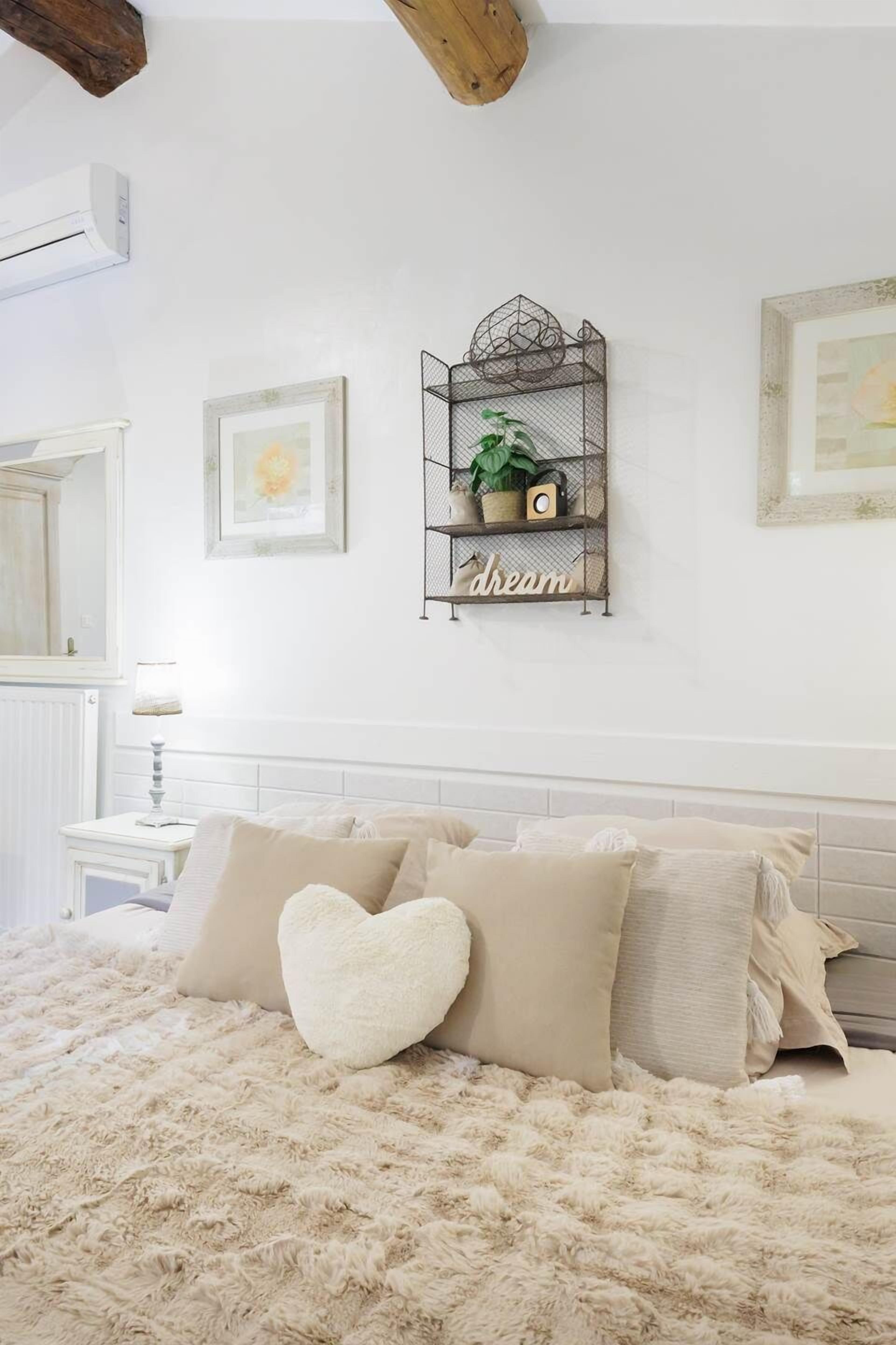 Een slaapkamer met een zacht beigekleurd bed, een hartvormig kussen, een metalen plank met een plant en een klok, en een lijst met een foto aan de muur.