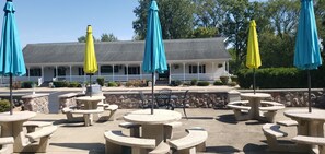 Outdoor seating with stone tables and blue and yellow umbrellas, a building with a porch, and a stone wall in the background.