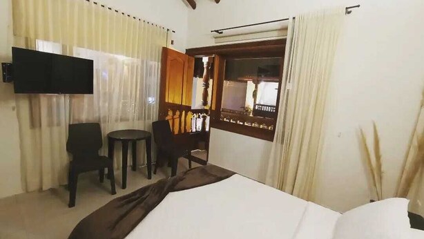 A hotel room with a bed, a TV mounted on the wall, a small table with chairs, and a window with curtains. - Hospedaje Villa Monica (Villa de Leyva)