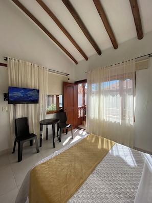 A hotel room with a bed, a TV, a desk, and a chair. There is a door leading to an outdoor area.