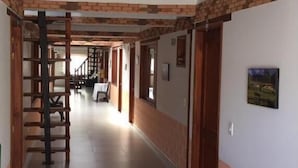 A hallway with a brick wall, wooden ceiling beams, and a staircase. There is a dining area with a table and chairs. - Hospedaje Villa Monica (Villa de Leyva)