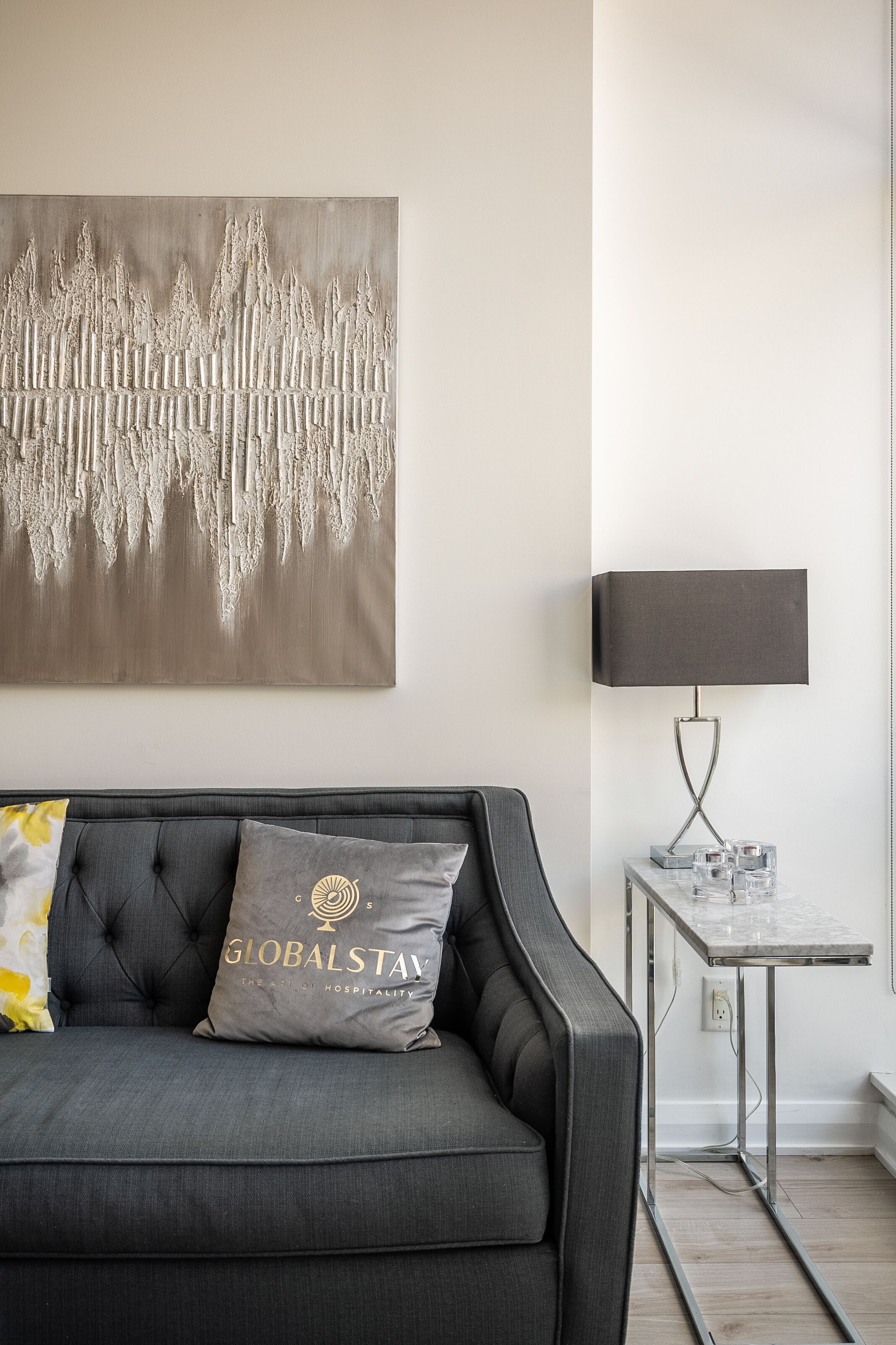 Ein modernes Wohnzimmer mit grauem Sofa, einem an der Wand befestigten abstrakten Kunstwerk, einem kleinen Beistelltisch mit Lampe und einem Zierkissen.