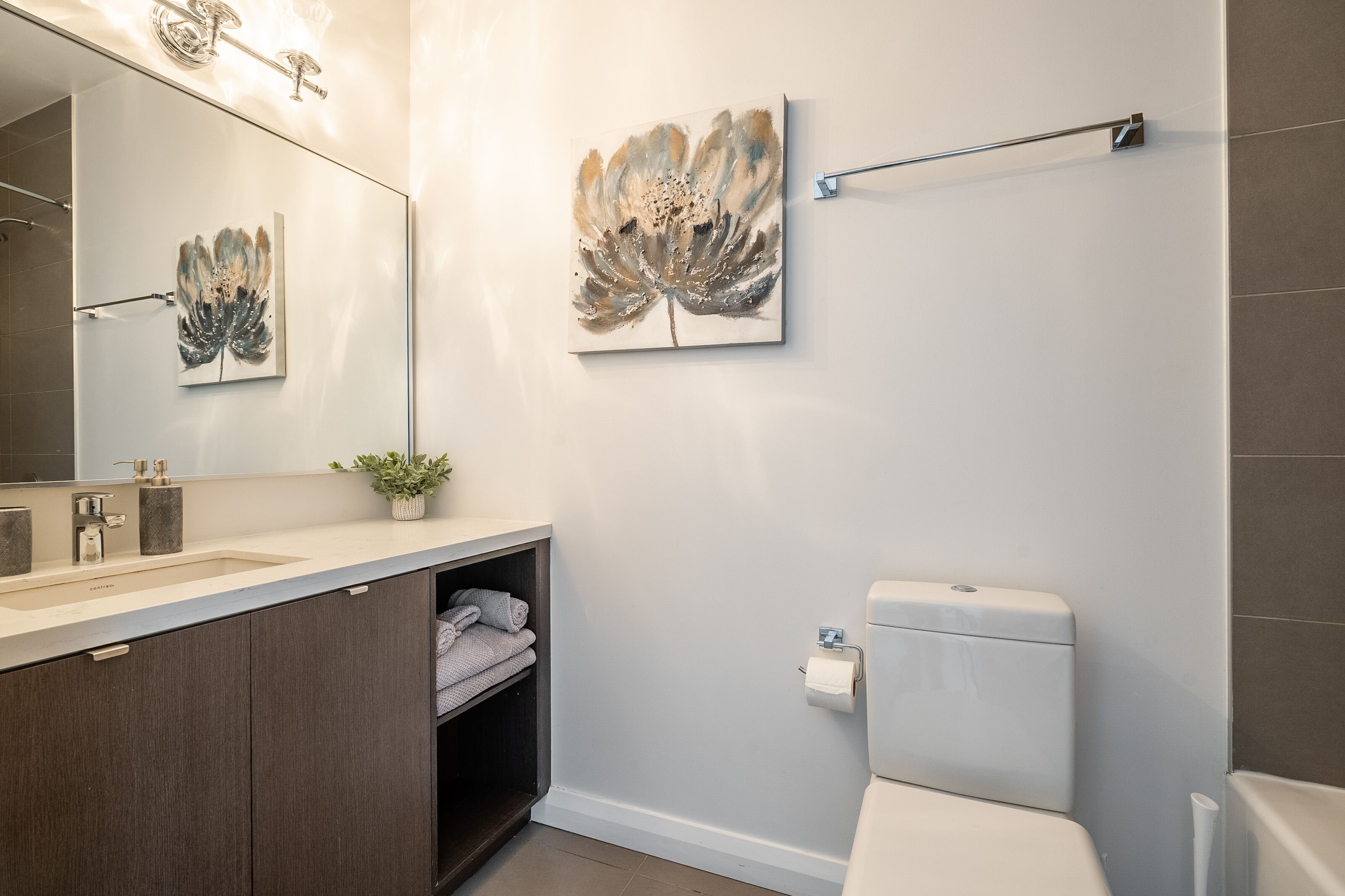 Un baño moderno con un espejo grande, un lavamanos con mueble oscuro, un inodoro y dos obras de arte colgadas en la pared.