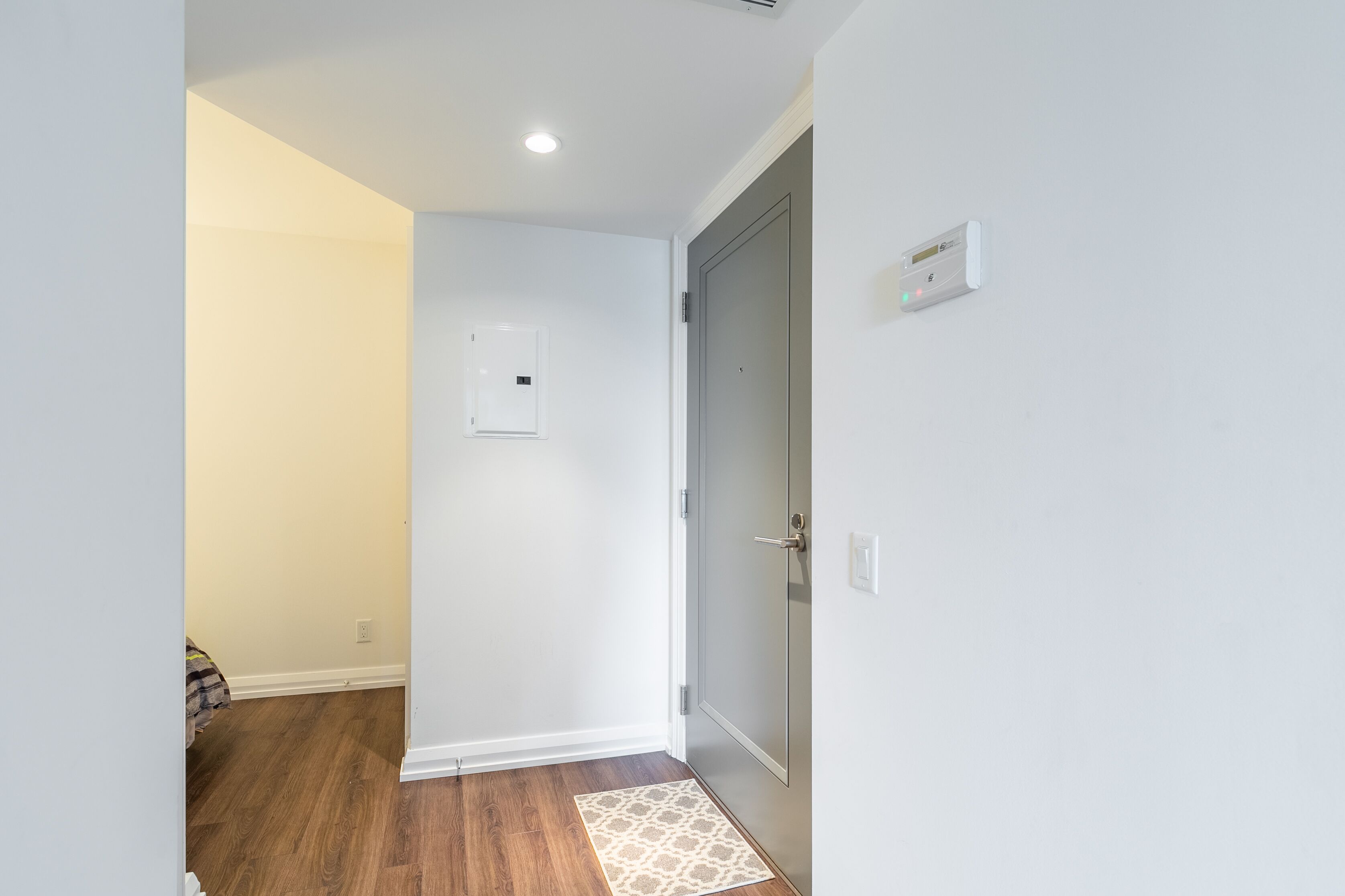 A hallway with a wooden floor, a white door with a lock, and a light switch on the wall.
