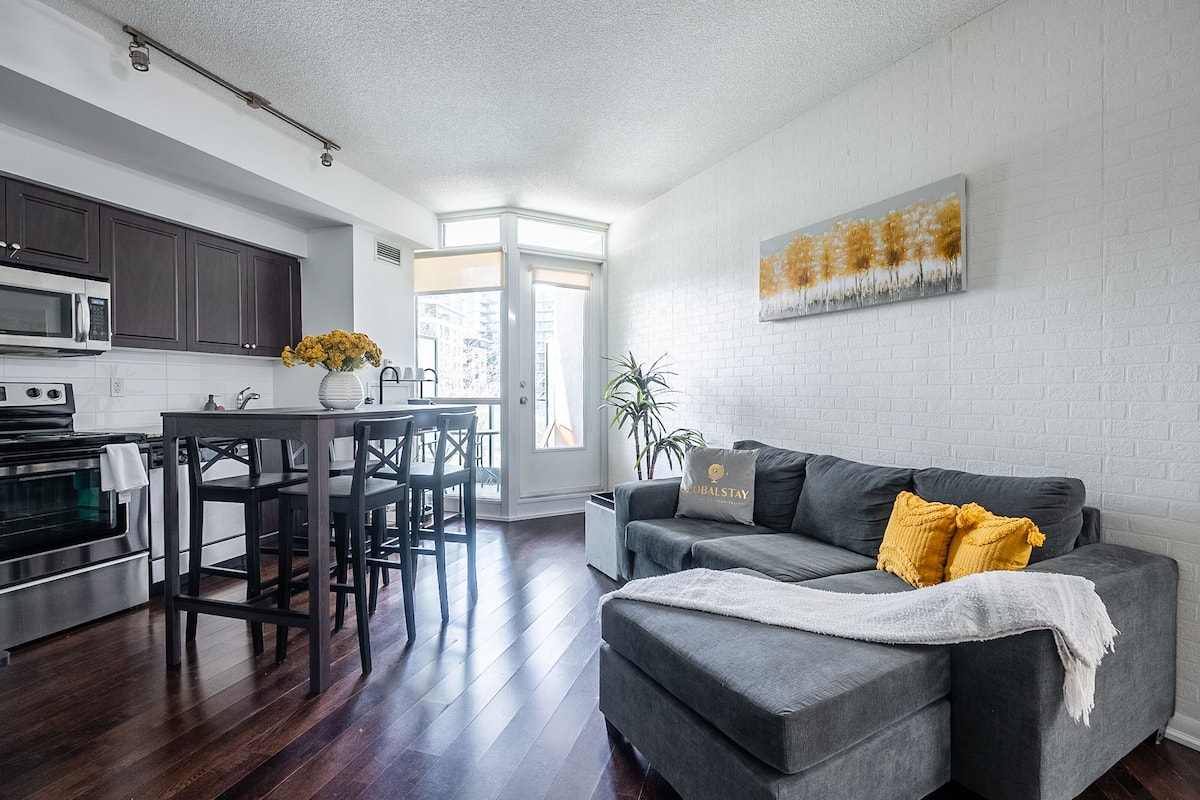 A modern kitchen with dark cabinets, a dining area with a table and chairs, a gray sofa with a yellow pillow, and a white brick wall with a landscape painting.