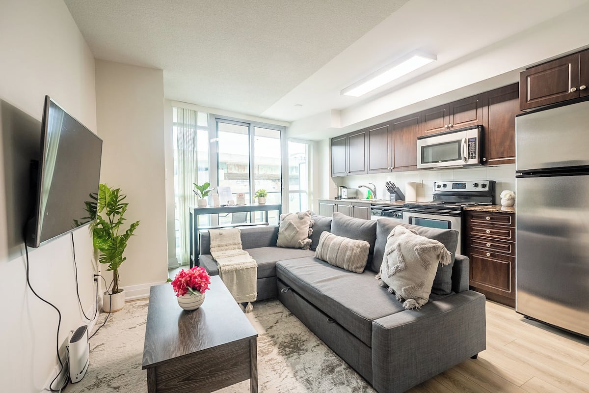 A modern living room with a grey sectional sofa, a flat-screen TV, a coffee table, and a kitchen area with a microwave, oven, and refrigerator.