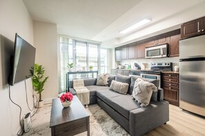 A modern living room with a grey sectional sofa, a flat-screen TV, a coffee table, and a kitchen area with a microwave, oven, and refrigerator. - Fort York Suites Toronto (Toronto)