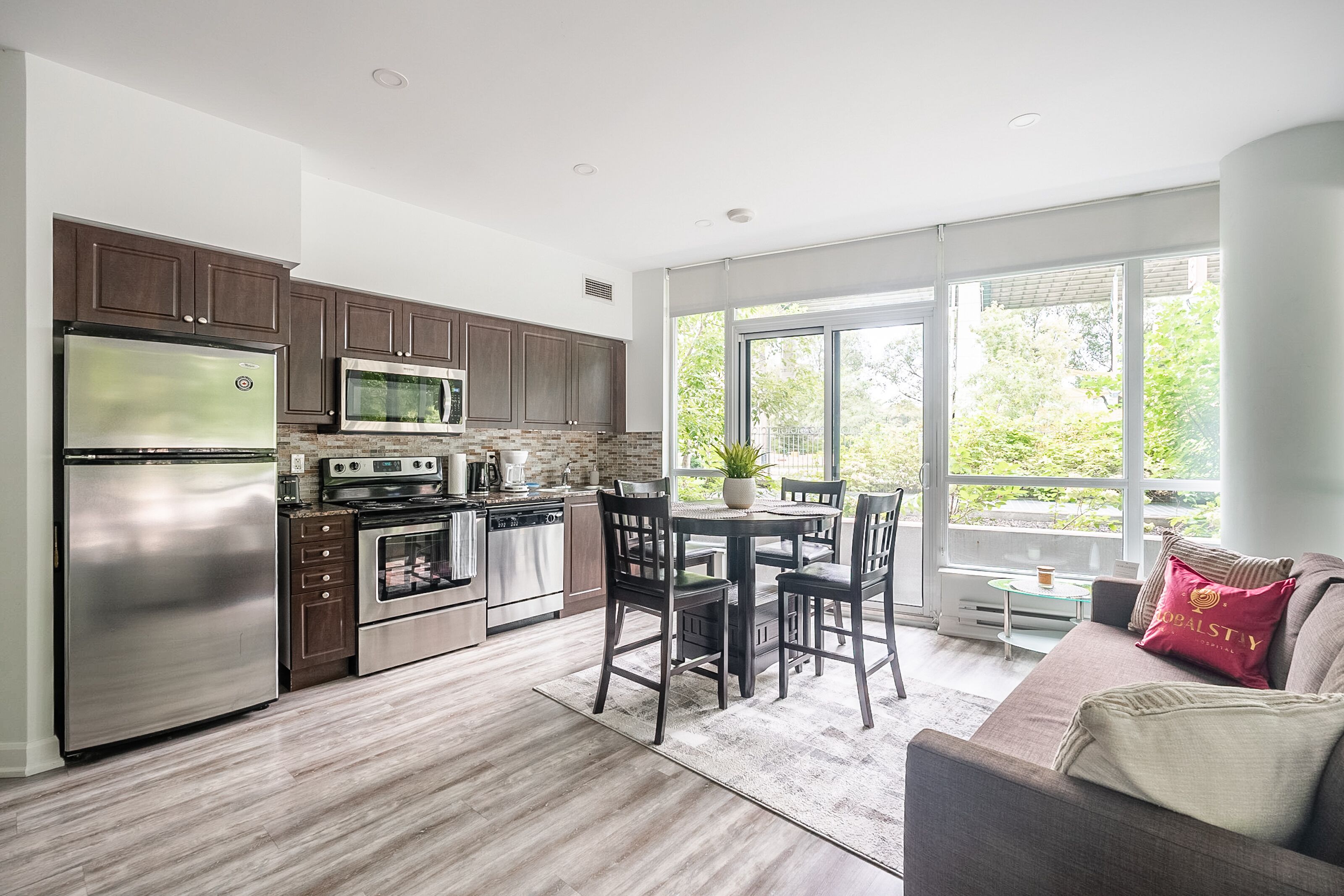 A modern kitchen with stainless steel appliances, a dining area with a table and chairs, and a large window offering a view of greenery.