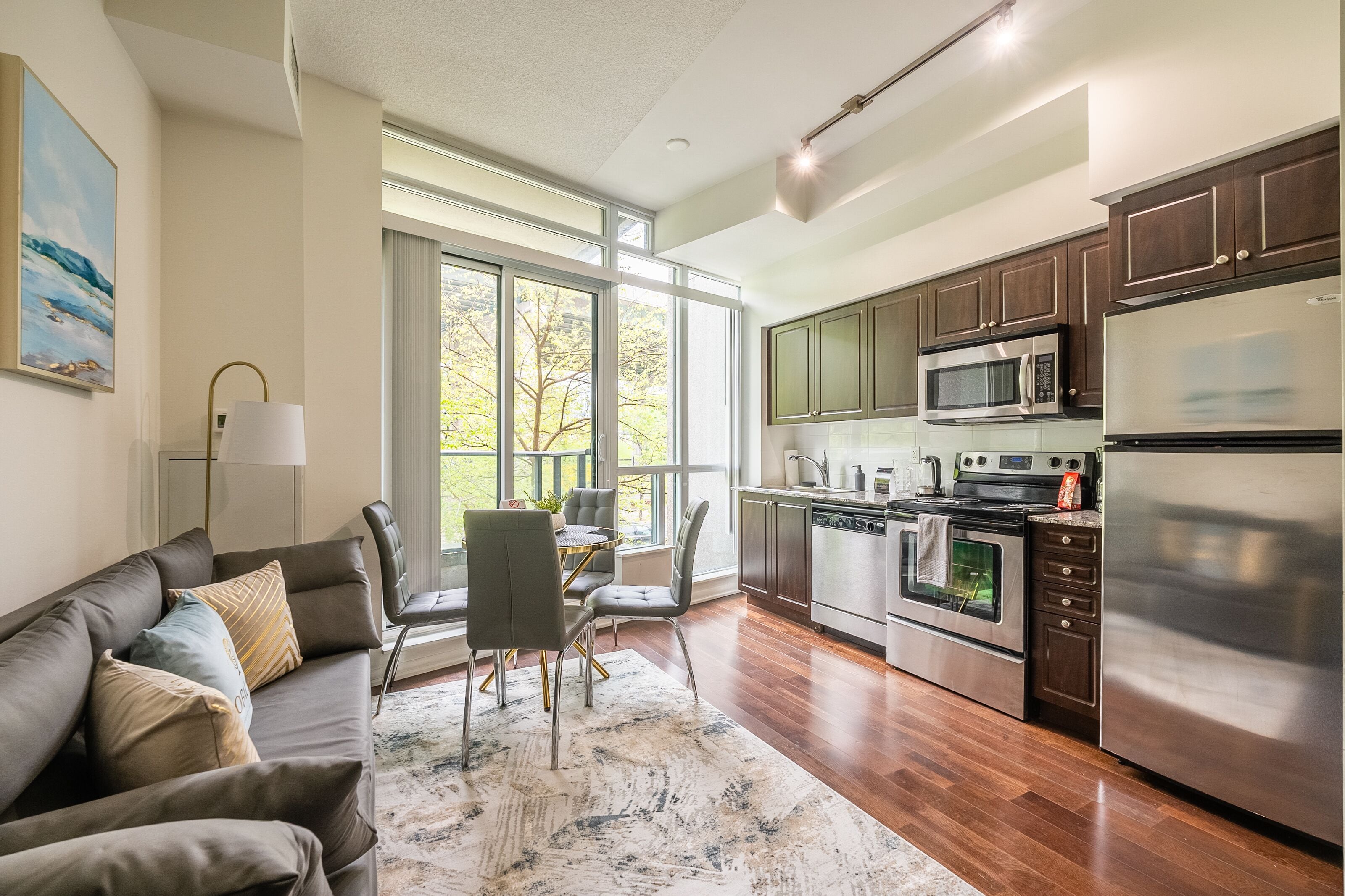 A modern kitchen with stainless steel appliances, a dining area with a gray couch and chairs, and large windows offering a view of trees.
