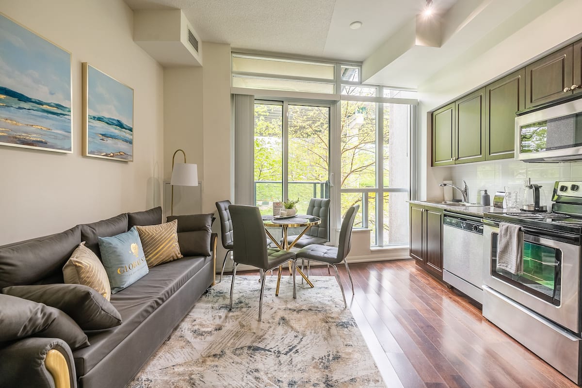 A modern kitchen with stainless steel appliances, a dining area with a round table and chairs, and a large window offering a view of trees.