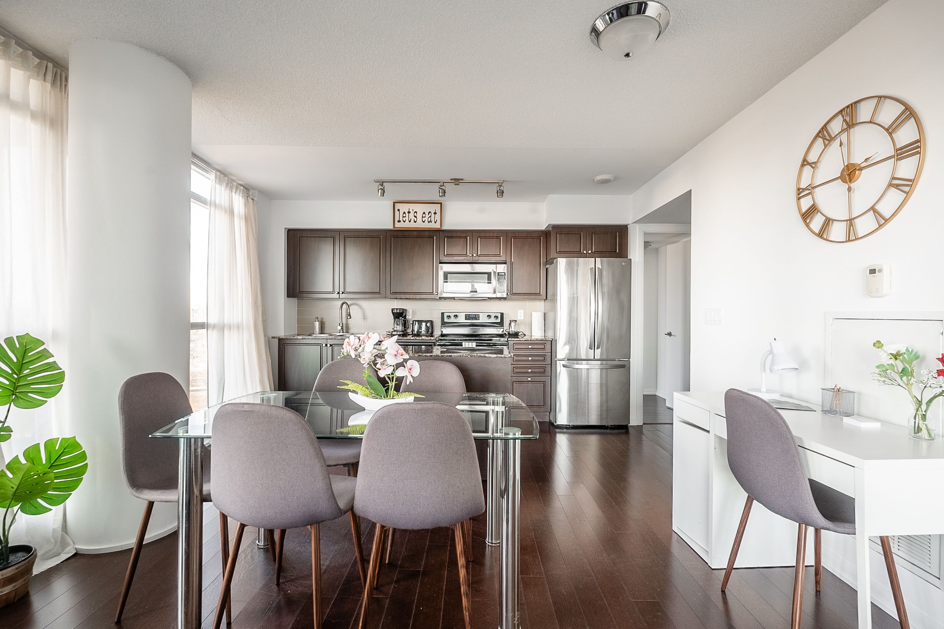 A modern kitchen with stainless steel appliances, a glass dining table, and a "Let's eat" sign.